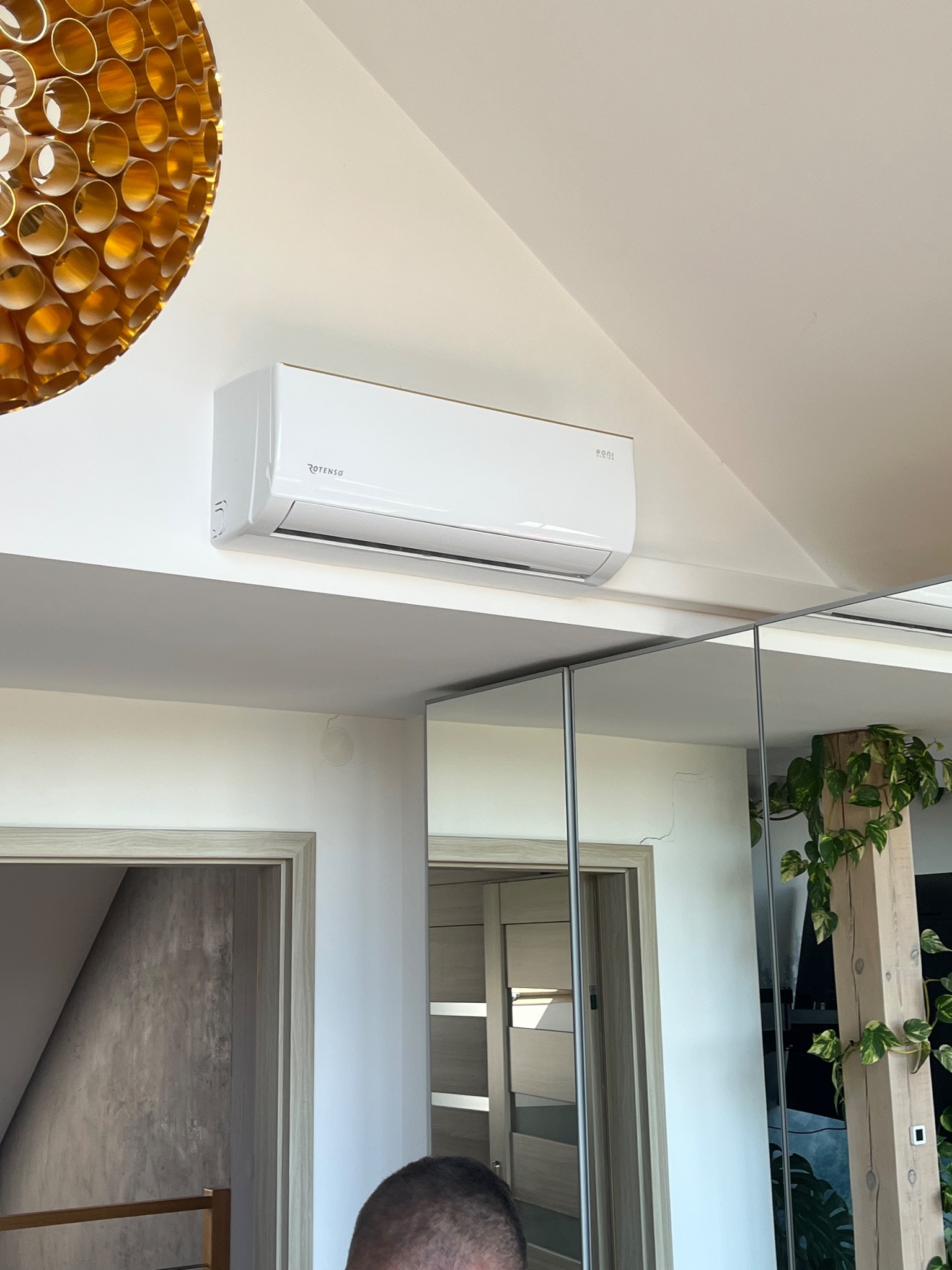 Biały klimatyzator Rotenso zamontowany na ścianie w jasnym wnętrzu, widoczny fragment lustra i dekoracyjna lampa sufitowa.