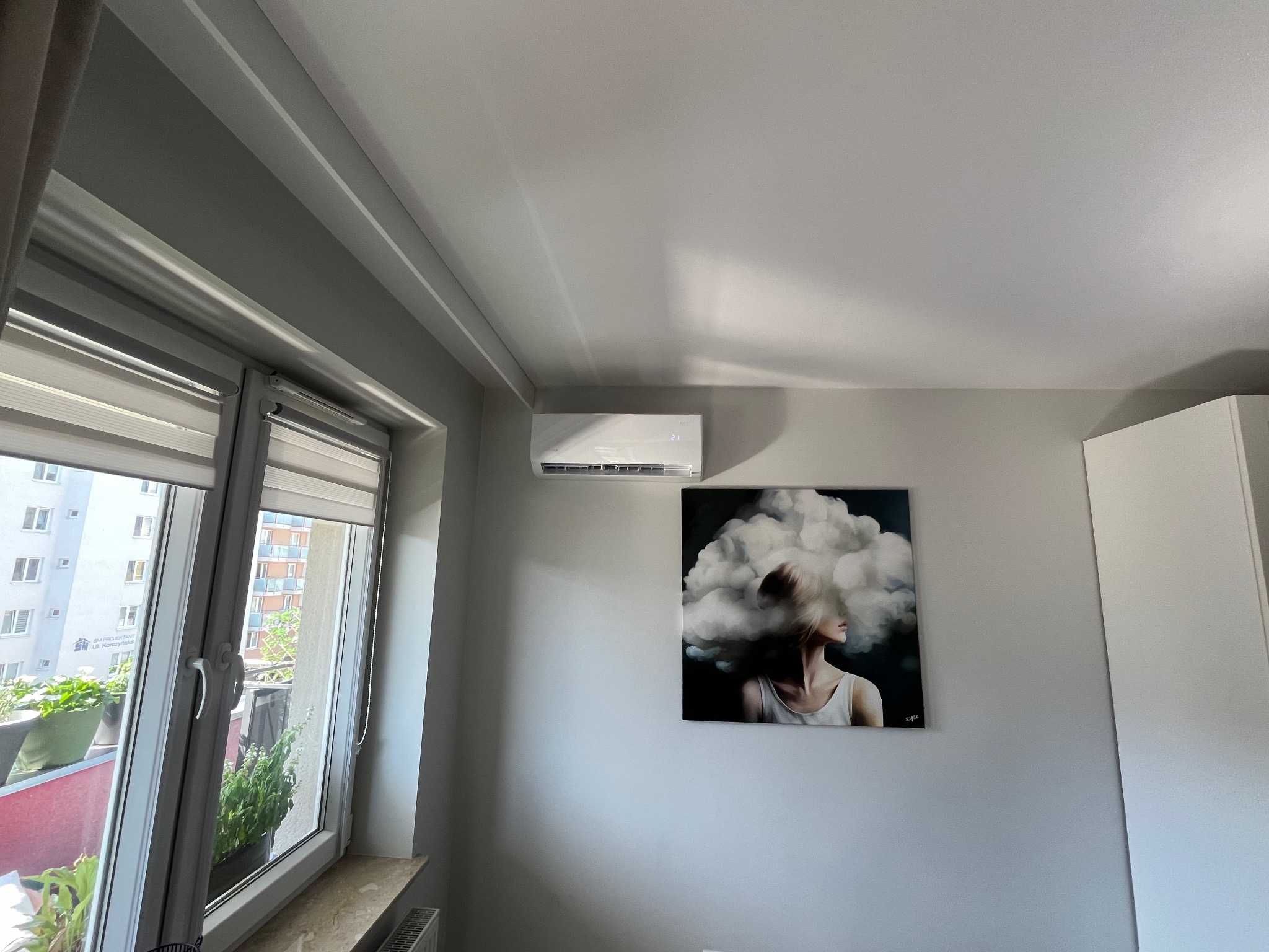 Biały klimatyzator ścienny zamontowany nad obrazem w nowoczesnym mieszkaniu z oknem balkonowym.