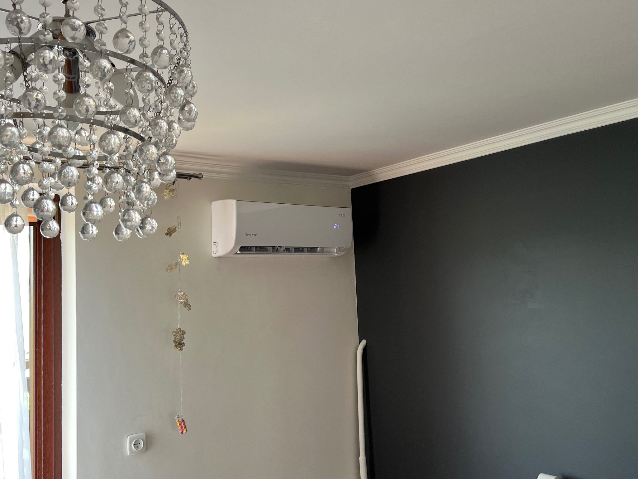 Biały klimatyzator Rotenso zamontowany na ścianie w pokoju z dekoracyjnym żyrandolem kryształowym, czarną ścianą i listwą przysufitową.