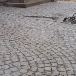 Ułożona z kostki granitowej nawierzchnia w odcieniach szarości, z widocznym fragmentem schodów i metalowym ogrodzeniem w tle. Fragment nawierzchni jest uszkodzony.