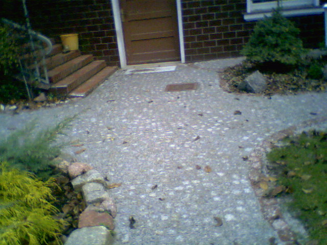 Chodnik wyłożony drobną kostką granitową prowadzący do drzwi wejściowych, obrzeża z kamieni, widoczne schody z cegły i roślinność.