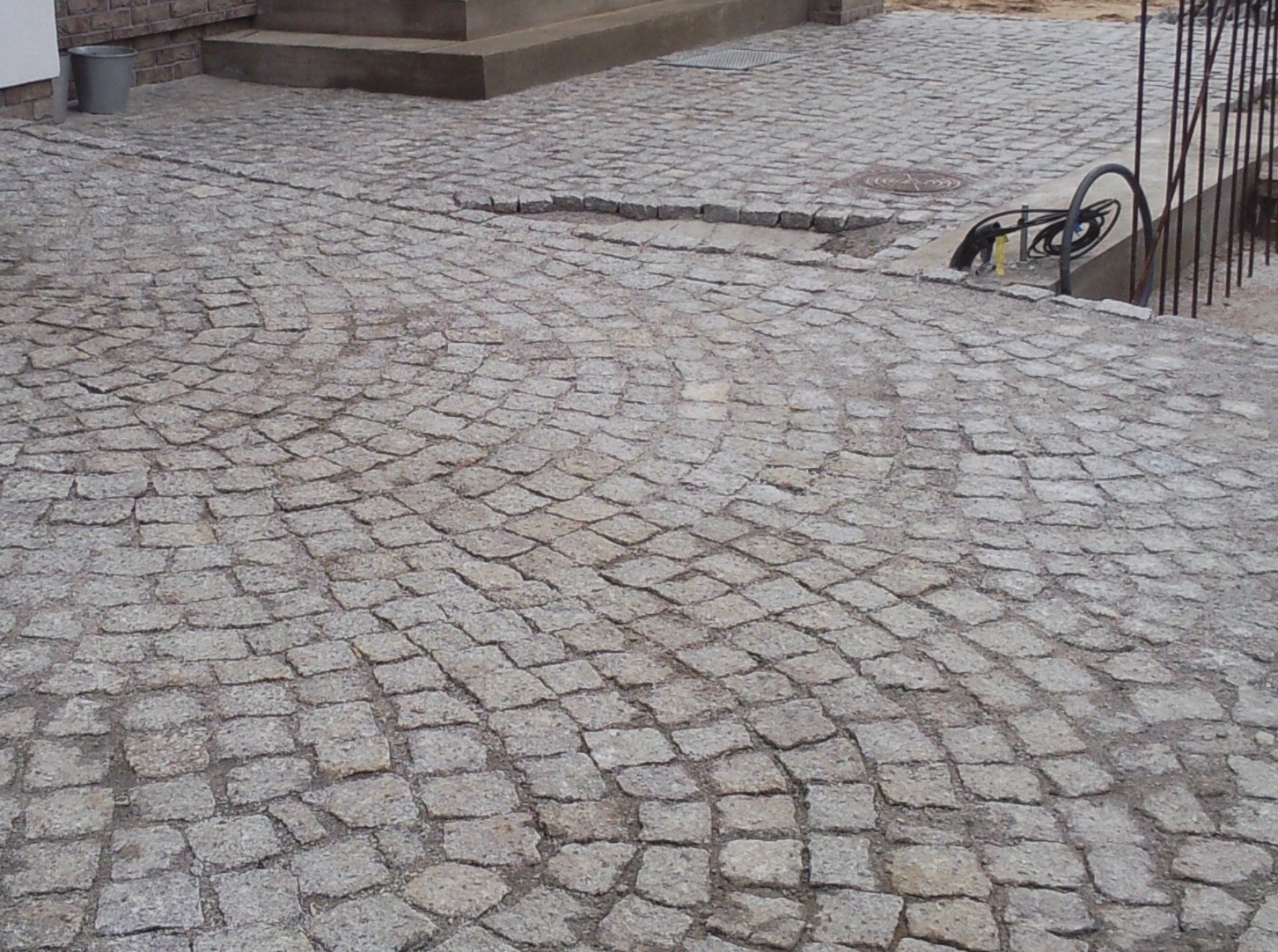 Ułożona z kostki granitowej nawierzchnia w odcieniach szarości, z widocznym fragmentem schodów i metalowym ogrodzeniem w tle. Fragment nawierzchni jest uszkodzony.