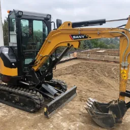 Żółto-czarna minikoparka SANY na placu budowy z widocznymi zbrojeniami w tle, pochmurne niebo.