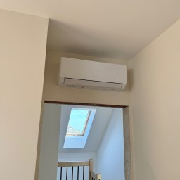 Kozak Klimatyzacja - Biały klimatyzator zamontowany na ścianie w domu, widoczny przez framugę drzwi, w tle schody i okno dachowe, instalator na drabinie.