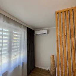 Biała klimatyzacja ścienna z wyświetlaczem temperatury, zamontowana w nowoczesnym salonie z drewnianą ścianką działową, zasłonami i lampą z wikliny.