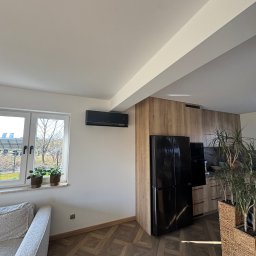 Kozak Klimatyzacja - Nowoczesny, czarny klimatyzator ścienny w jasnym wnętrzu z drewnianą podłogą i zabudową kuchenną. Widok na okno z roślinami i fragment sofy.