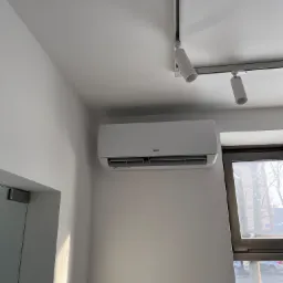 Biały klimatyzator AUX zamontowany na ścianie w jasnym pomieszczeniu, widok z boku, oświetlenie punktowe na suficie, okno z widokiem na zimowy krajobraz.