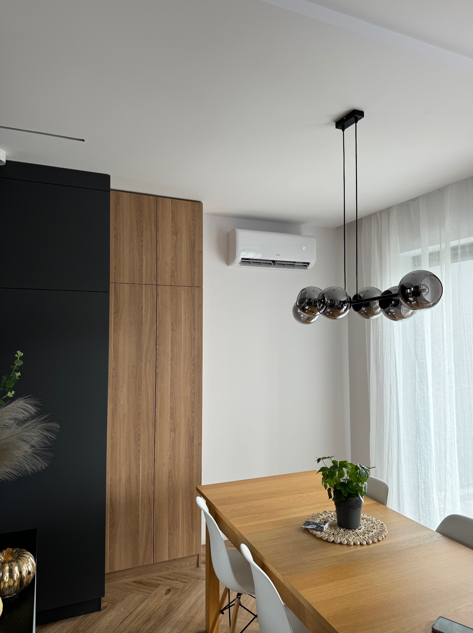 Nowoczesne wnętrze jadalni z klimatyzatorem ściennym, wiszącą lampą z dymionego szkła i drewnianym stołem z rośliną w doniczce.