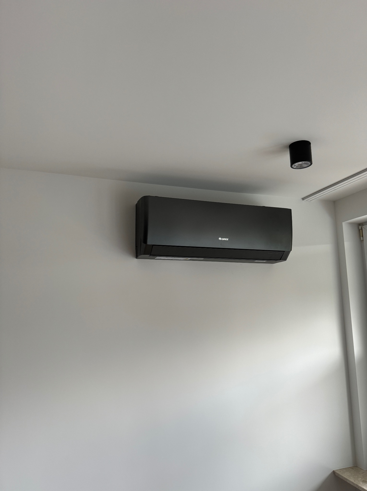 Czarny klimatyzator marki Gree zamontowany na białej ścianie w pomieszczeniu z czarną lampą sufitową.