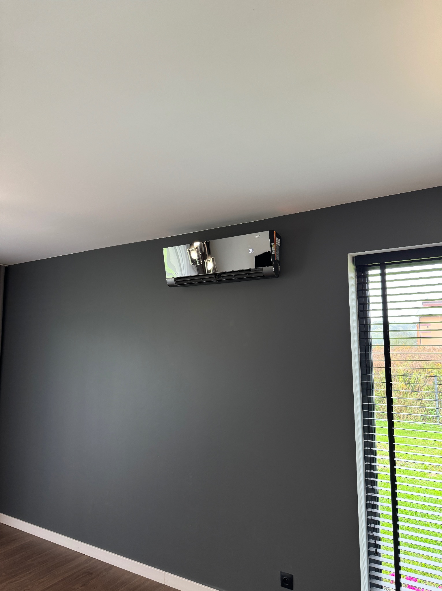 Czarny klimatyzator ścienny z wyświetlaczem temperatury, zamontowany w nowoczesnym wnętrzu z ciemnoszarą ścianą, białym sufitem i drewnianą podłogą, widok z okna z żaluzjami.