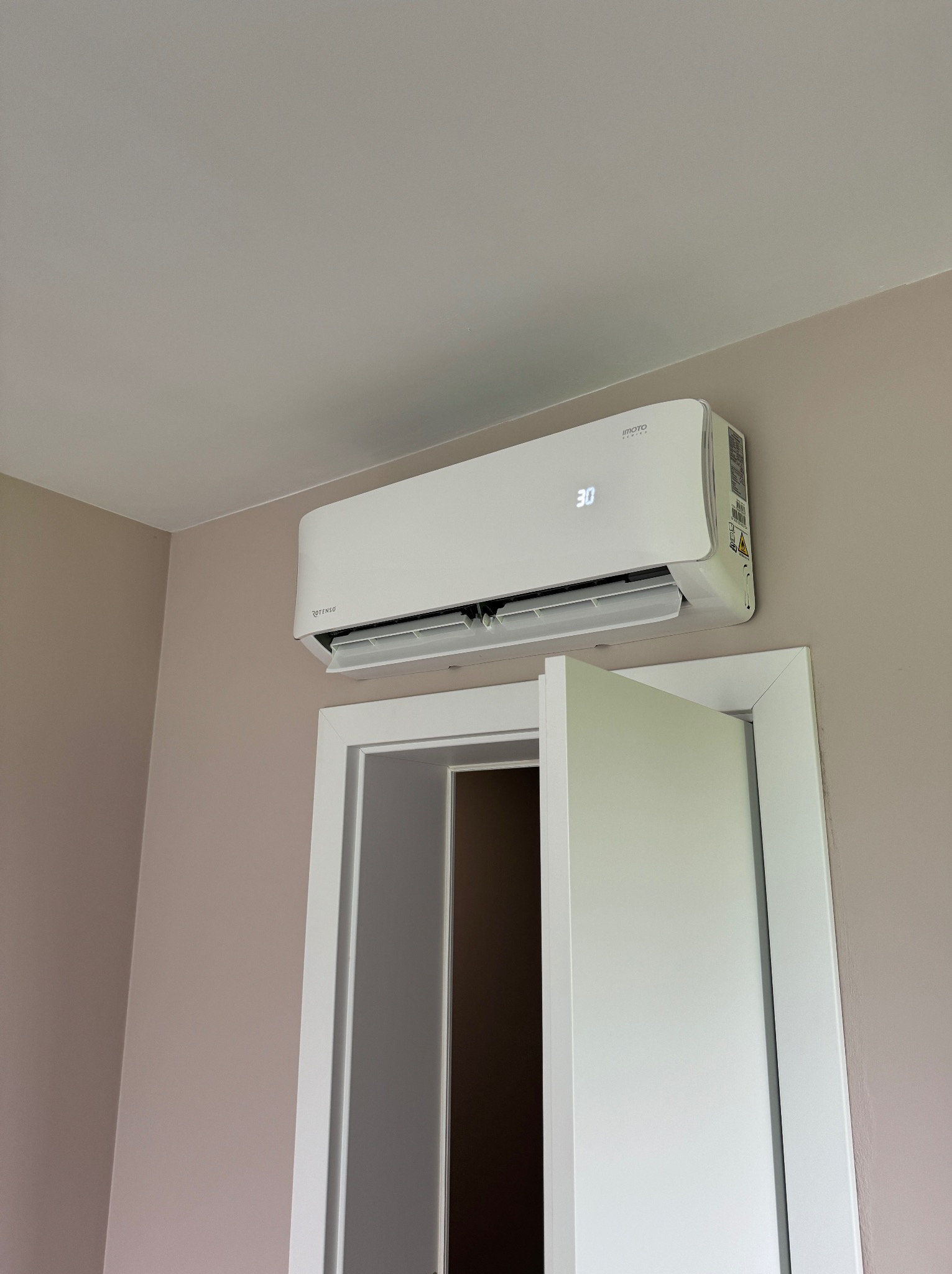 Biały klimatyzator ścienny z wyświetlaczem temperatury 30 stopni, zamontowany nad otwartymi, białymi drzwiami w pokoju o ścianach w kolorze beżowym.