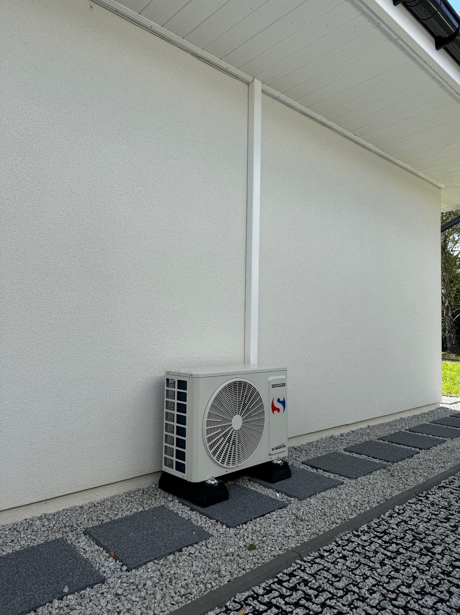 Zewnętrzna jednostka klimatyzacji Sinclair zamontowana przy białej ścianie domu, na czarnych podkładkach, z widoczną ścieżką z szarych płyt i kamieni.