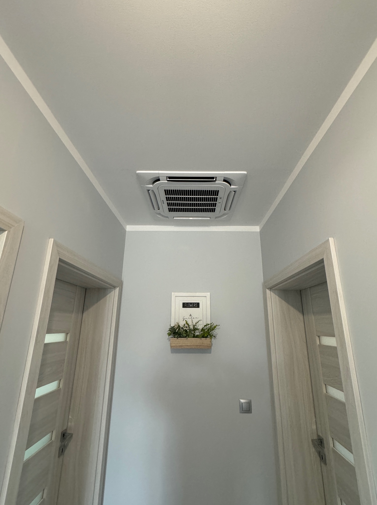 Kasetonowa klimatyzacja sufitowa w jasnym pomieszczeniu z dwoma drzwiami i dekoracją ścienną z roślinami.