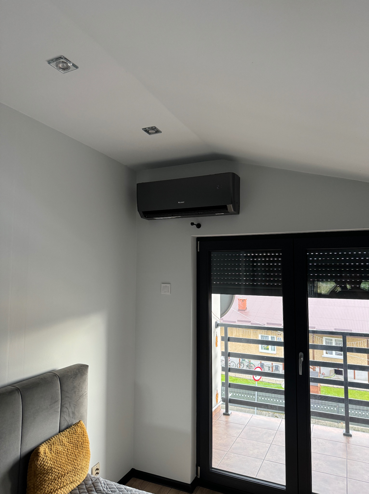 Czarny klimatyzator marki Gree zamontowany na ścianie nad czarnym oknem balkonowym z roletami w nowoczesnym pokoju z szarą tapicerowaną ramą łóżka i żółtą poduszką.