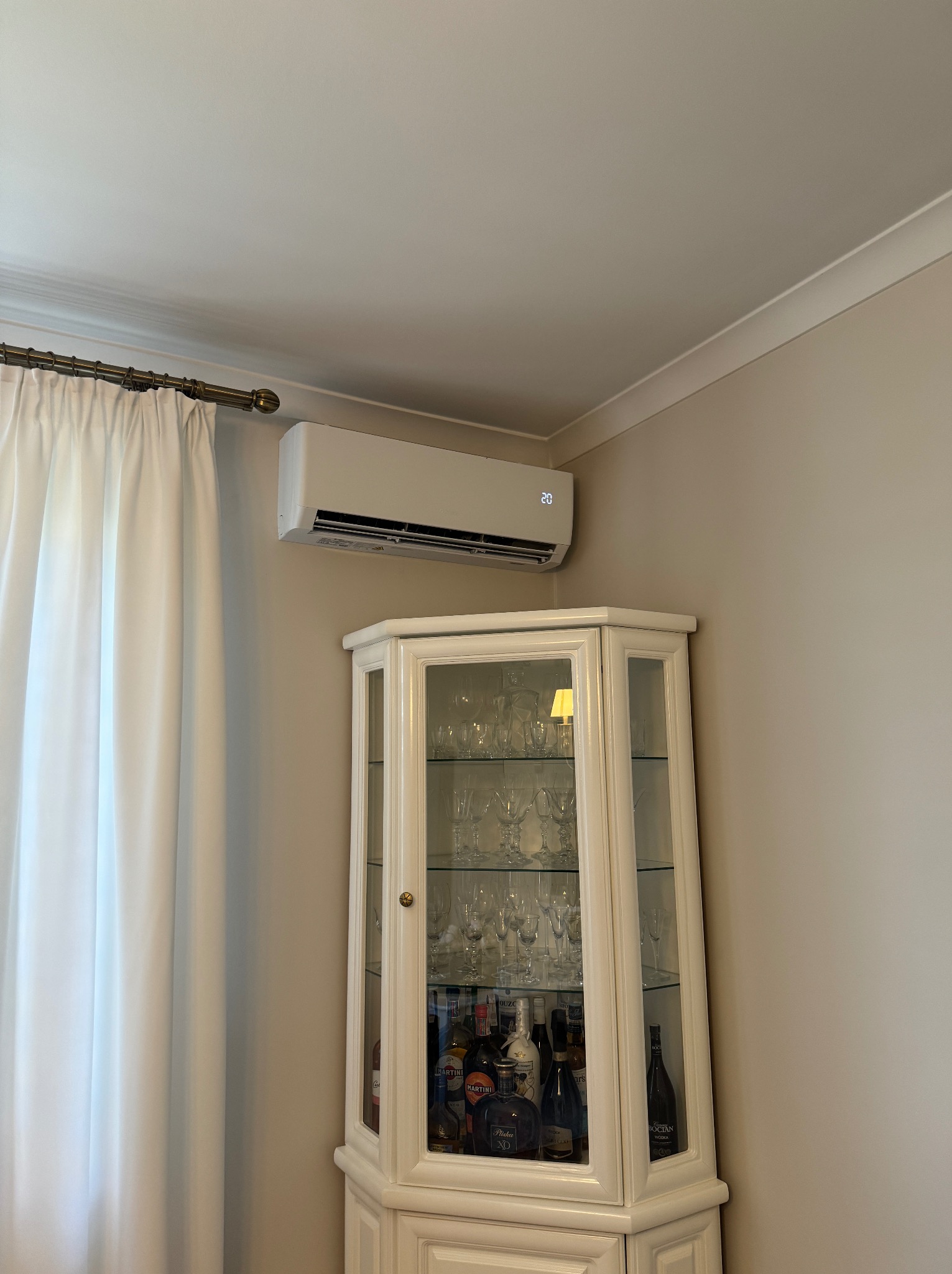 Klimatyzator ścienny zamontowany w salonie nad białą witryną z kieliszkami i butelkami alkoholu, obok zasłoniętego okna.