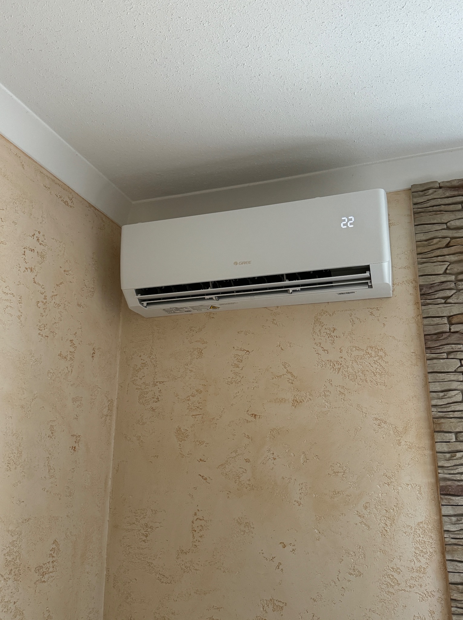 Biały klimatyzator marki Gree zamontowany na ścianie o beżowej, teksturowanej powierzchni, z widocznym wyświetlaczem temperatury ustawionym na 22 stopnie.