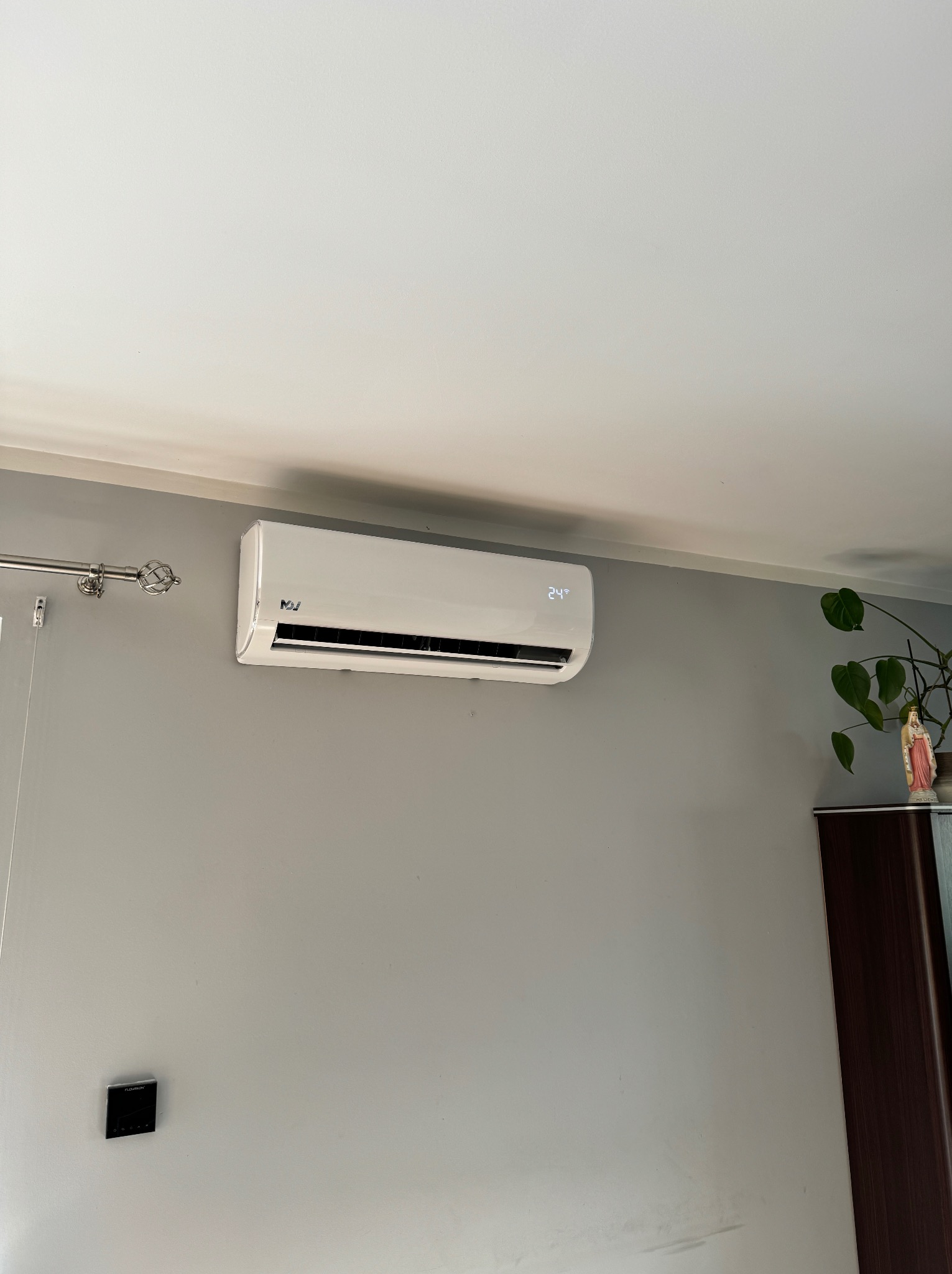 Biała klimatyzacja ścienna z wyświetlaczem temperatury 24 stopnie Celsjusza, zamontowana w szarym pokoju obok karnisza i szafy z rośliną doniczkową.