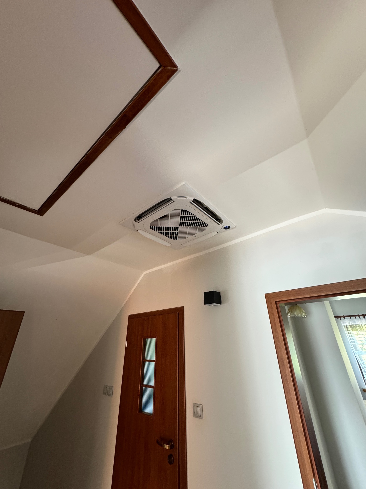 Kwadratowa jednostka klimatyzacyjna zamontowana w suficie poddasza, widoczna z dołu, z drewnianymi detalami wykończeniowymi.