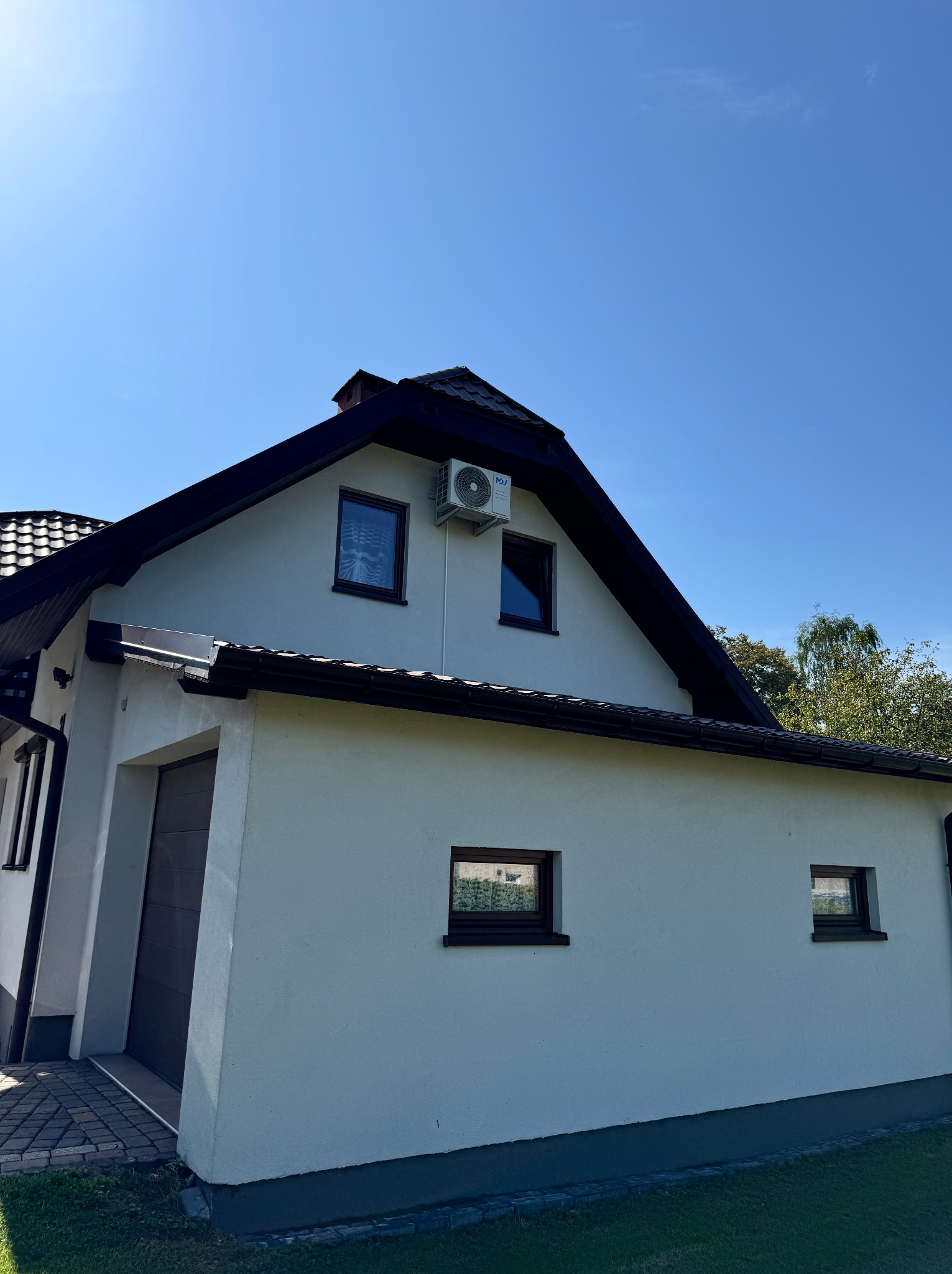 Biały dom jednorodzinny z ciemnobrązowym dachem i zamontowaną jednostką klimatyzacji zewnętrznej pod oknem na poddaszu, widok z dołu na tle błękitnego nieba.