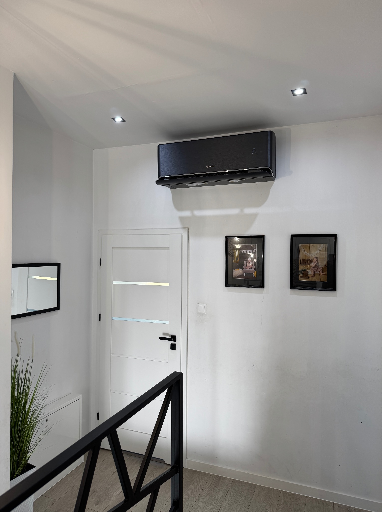 Czarny klimatyzator zamontowany na białej ścianie w nowoczesnym wnętrzu, widoczne zdjęcia w ramkach, białe drzwi i fragment czarnej balustrady schodów.
