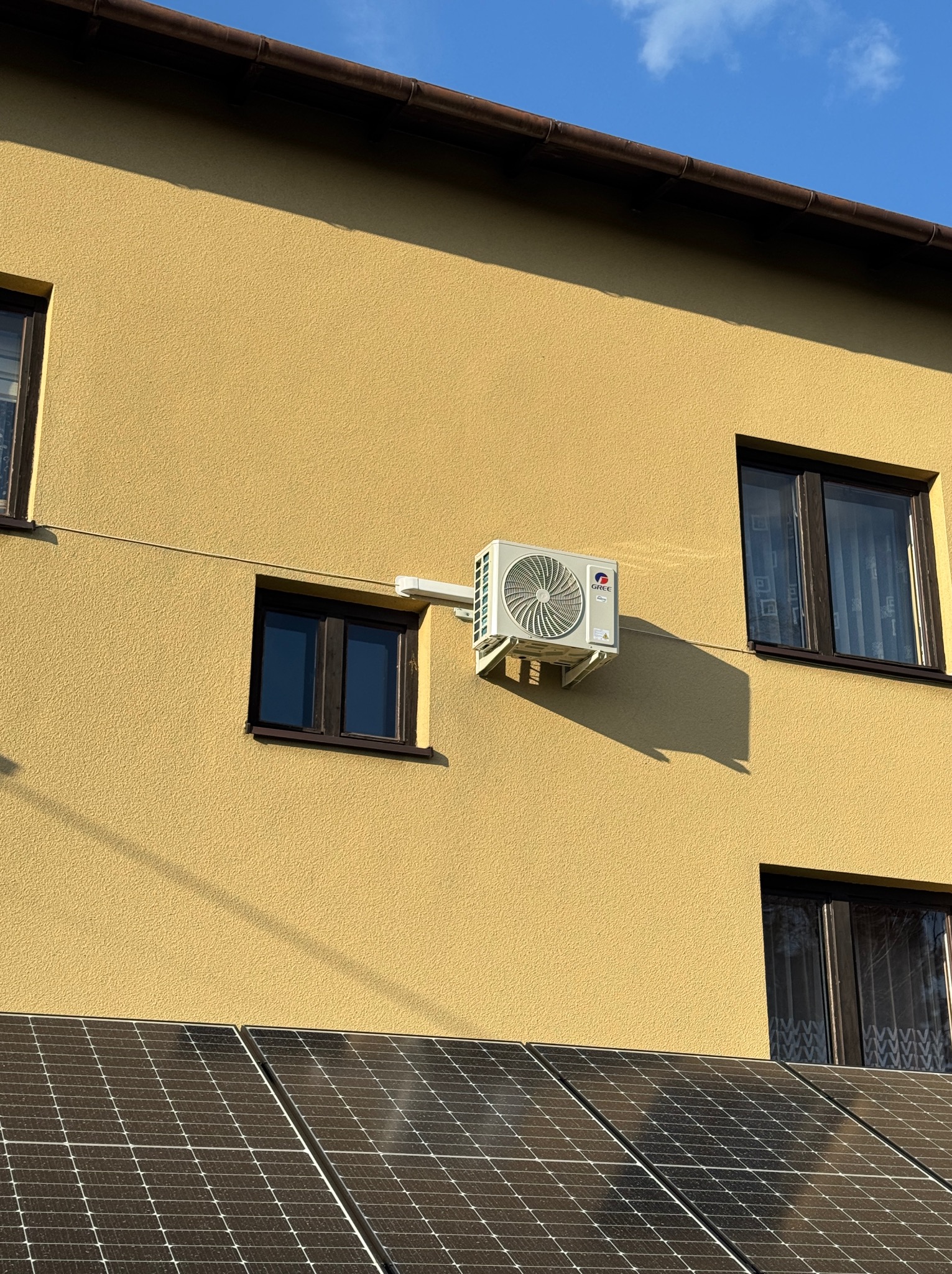 Zewnętrzna jednostka klimatyzacji marki Gree zamontowana na żółtej elewacji domu, widoczne panele słoneczne na dachu.