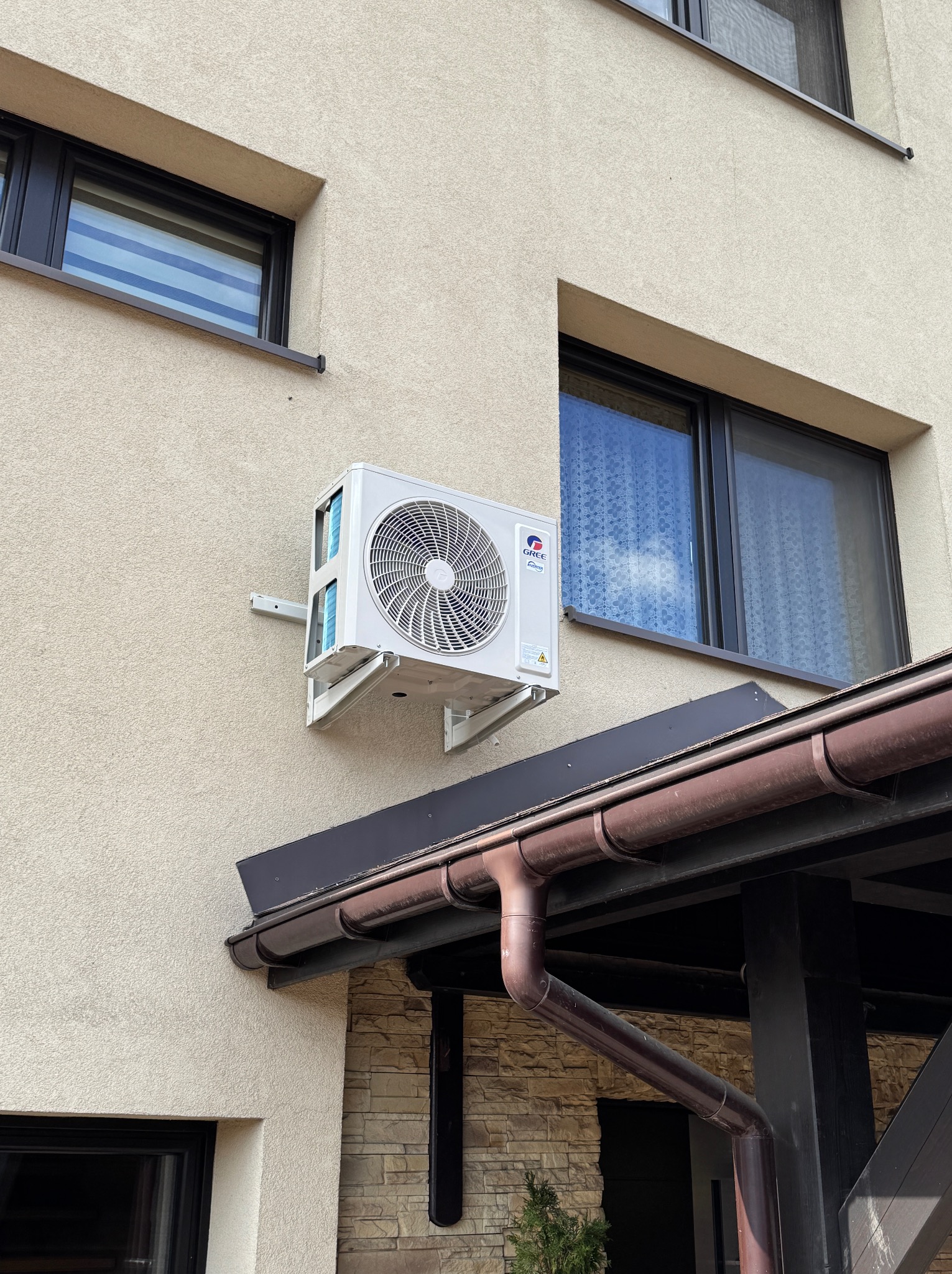 Biała jednostka zewnętrzna klimatyzatora Gree zamontowana na kremowej elewacji domu, widoczne dwa okna z czarnymi ramami i brązowa rynna.
