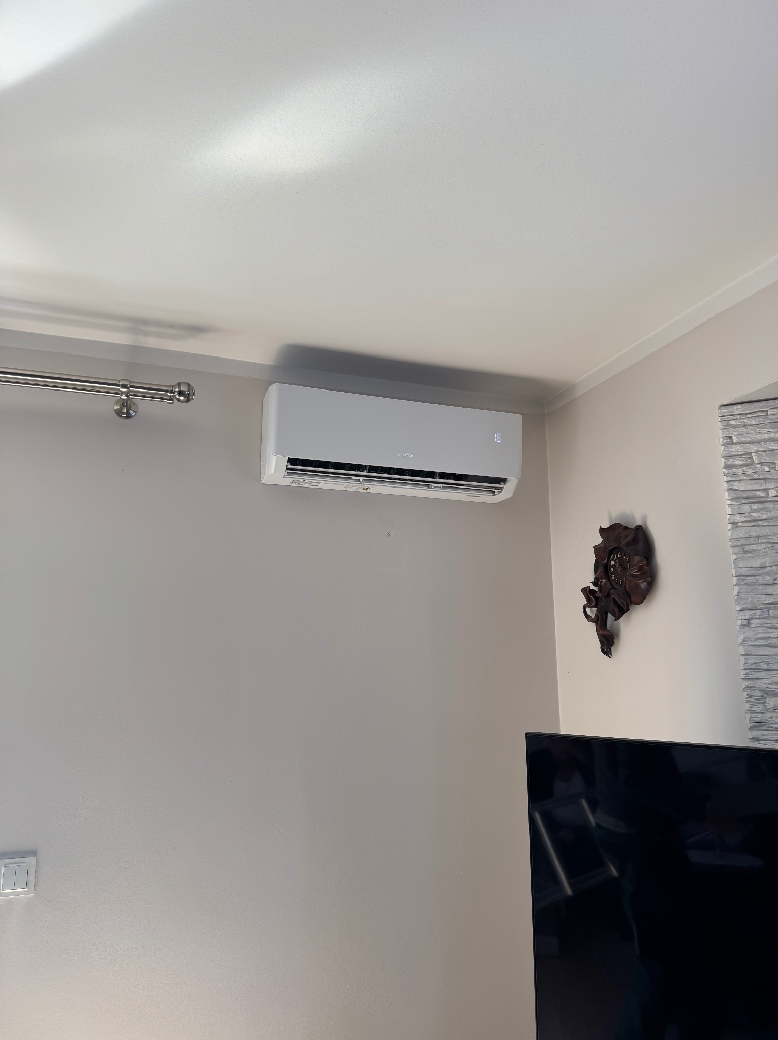 Biały klimatyzator marki Gree zamontowany na ścianie w pokoju, wyświetlający temperaturę 16 stopni Celsjusza, obok dekoracyjny zegar i fragment ściany wykończonej imitacją kamienia.