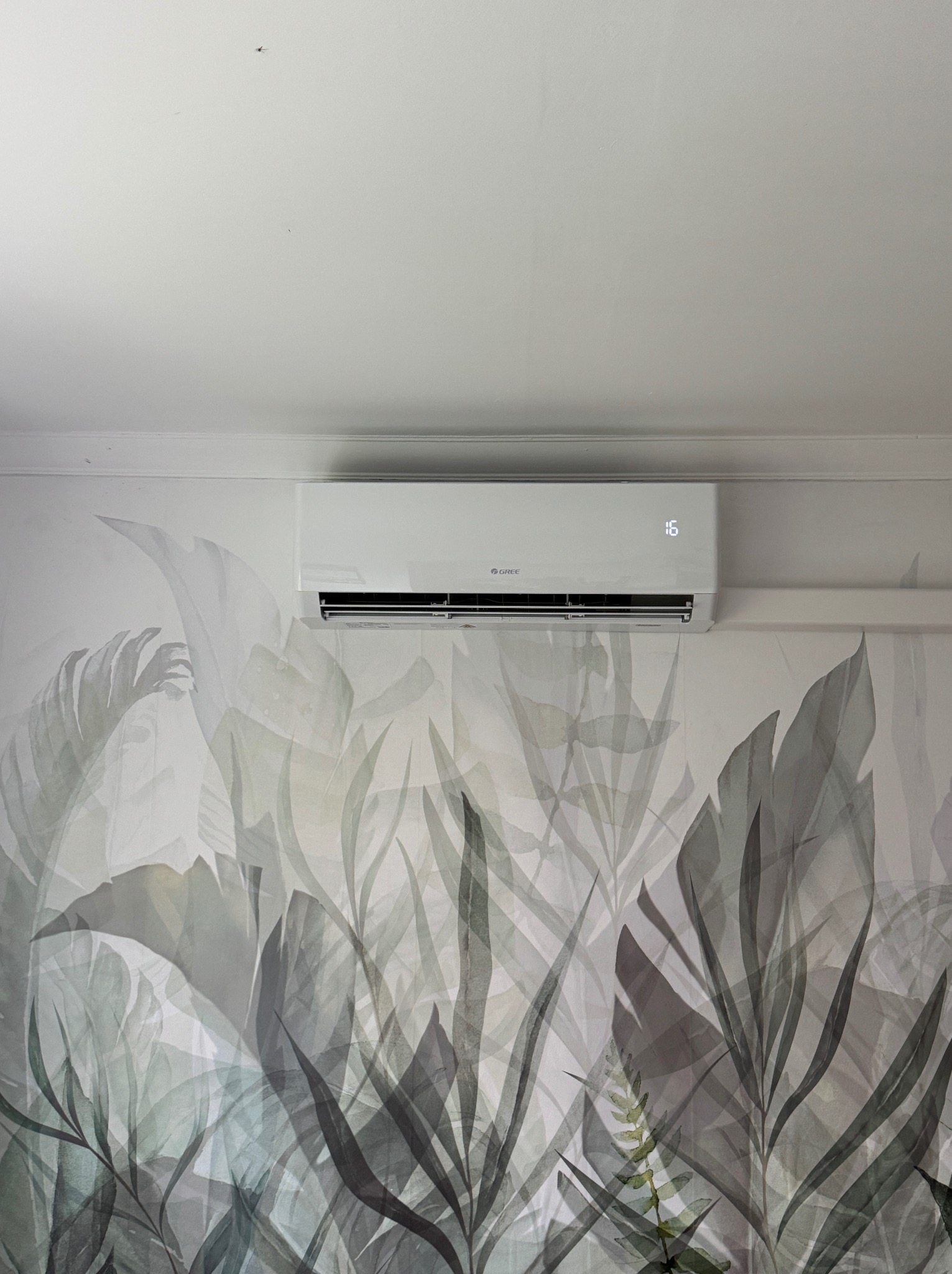 Nowoczesny klimatyzator marki Gree zamontowany na ścianie z dekoracyjną tapetą w motywy roślinne.