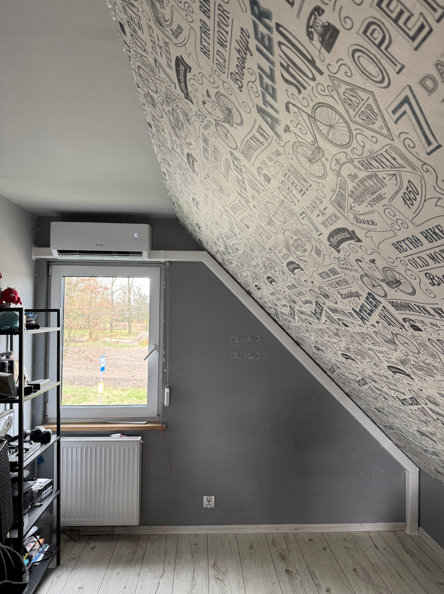 Klimatyzator zamontowany w pokoju na poddaszu ze skośnym sufitem pokrytym tapetą w stylu retro z motywami rowerów i napisów.