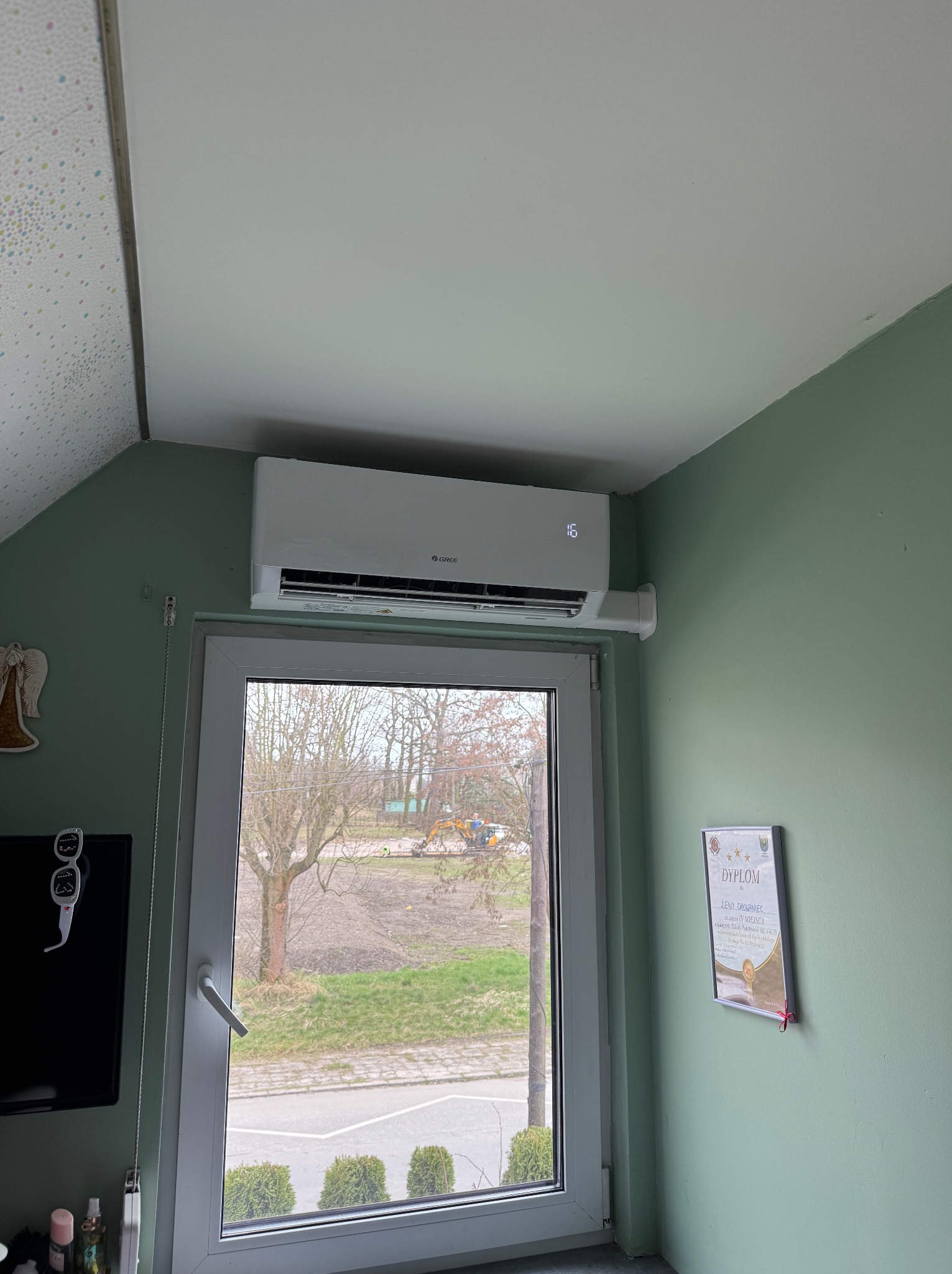 Biały klimatyzator marki Gree zamontowany nad oknem w pokoju o zielonych ścianach, widoczny wyświetlacz temperatury ustawionej na 16 stopni Celsjusza oraz dyplom wiszący na ścianie.