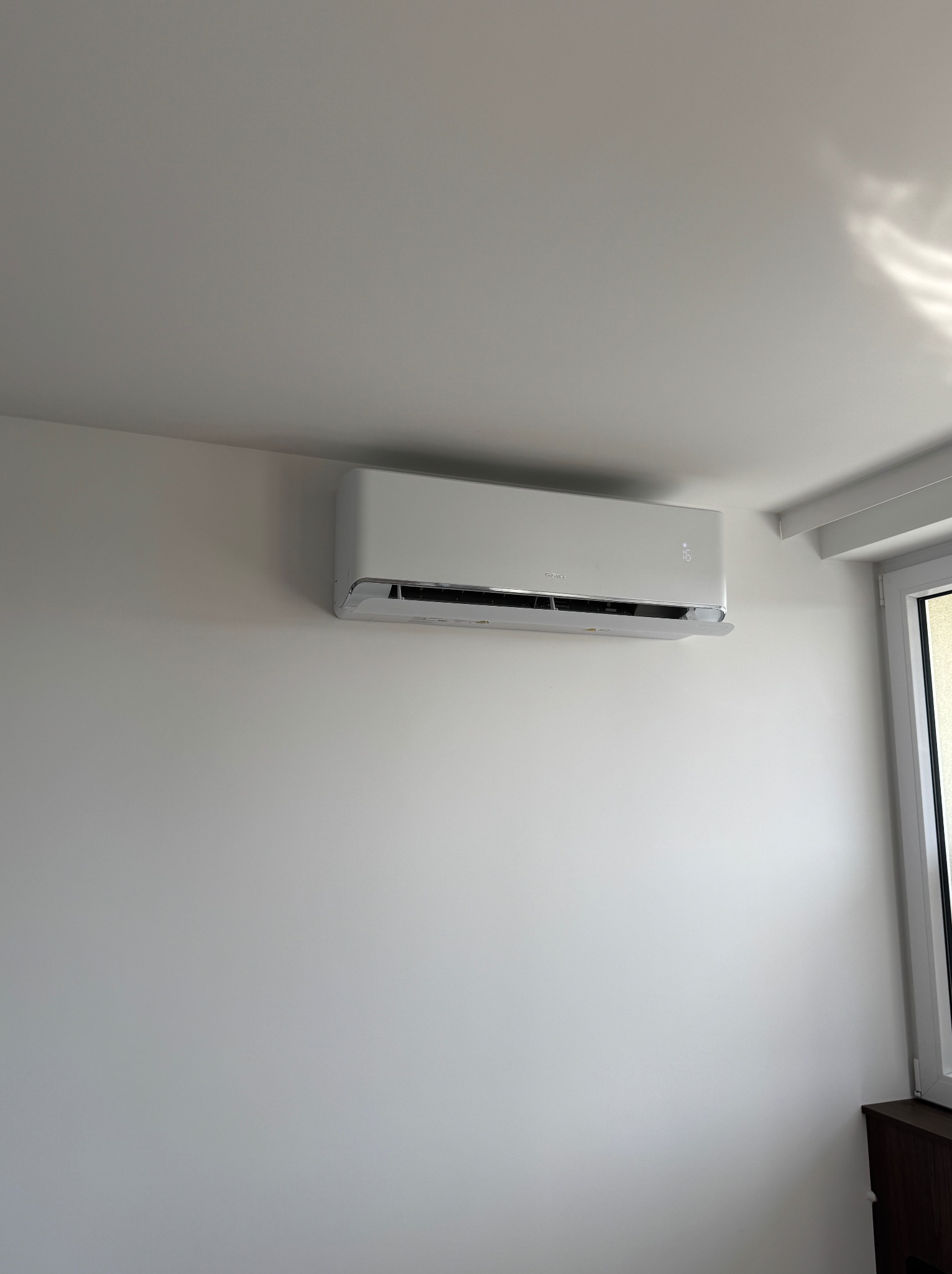 Biała, nowoczesna klimatyzacja ścienna marki Gree zamontowana w jasnym pomieszczeniu z oknem.