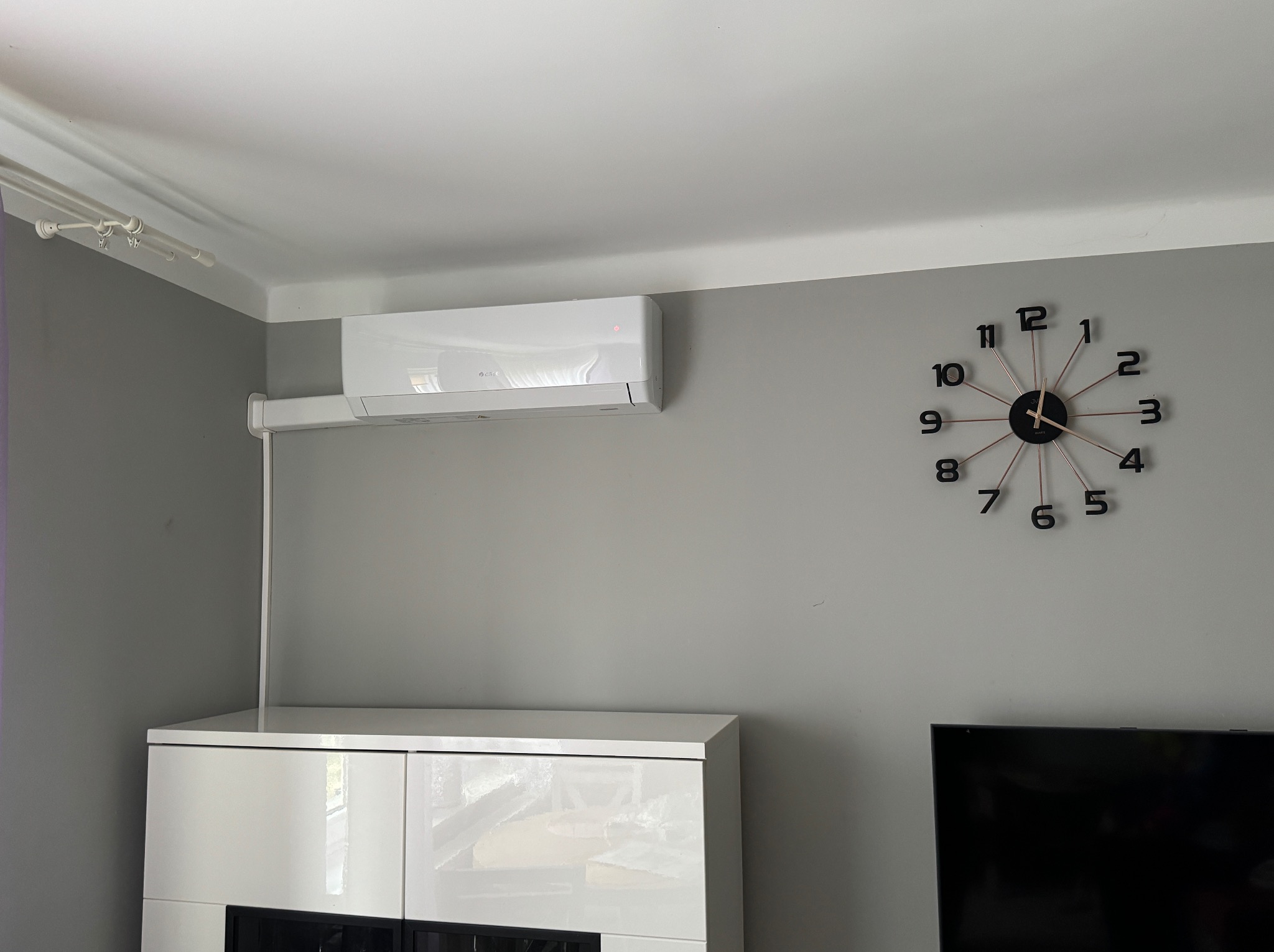 Biały klimatyzator ścienny marki Gree zamontowany w szarym salonie nad białą komodą, obok zegara ściennego.