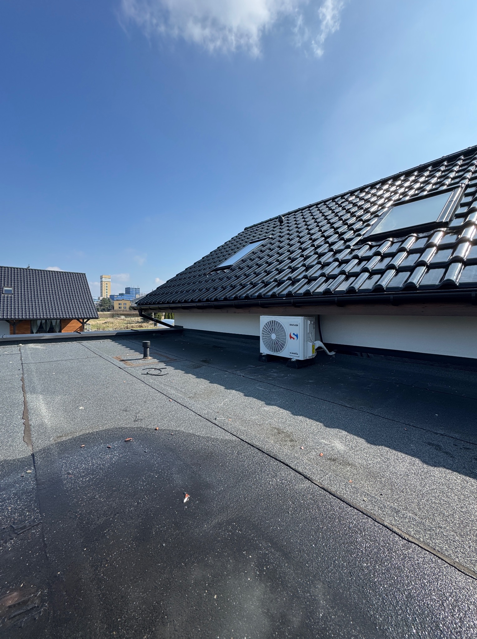 Biała jednostka zewnętrzna klimatyzacji zamontowana na płaskim dachu domu jednorodzinnego, widok z góry na dach z czarną dachówką i oknami dachowymi, błękitne niebo w tle.