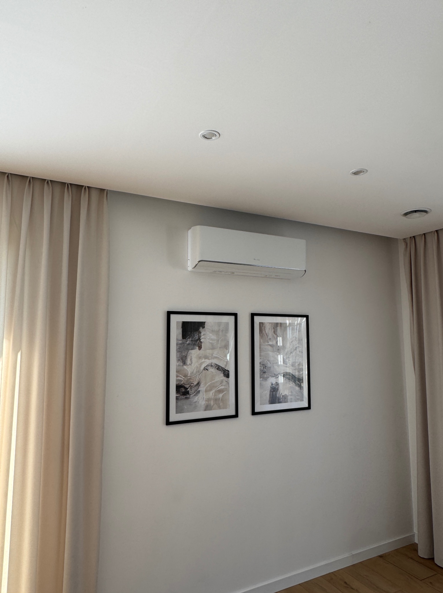 Biała jednostka klimatyzacji zamontowana na ścianie w pokoju z zasłonami i obrazami w czarnych ramkach.