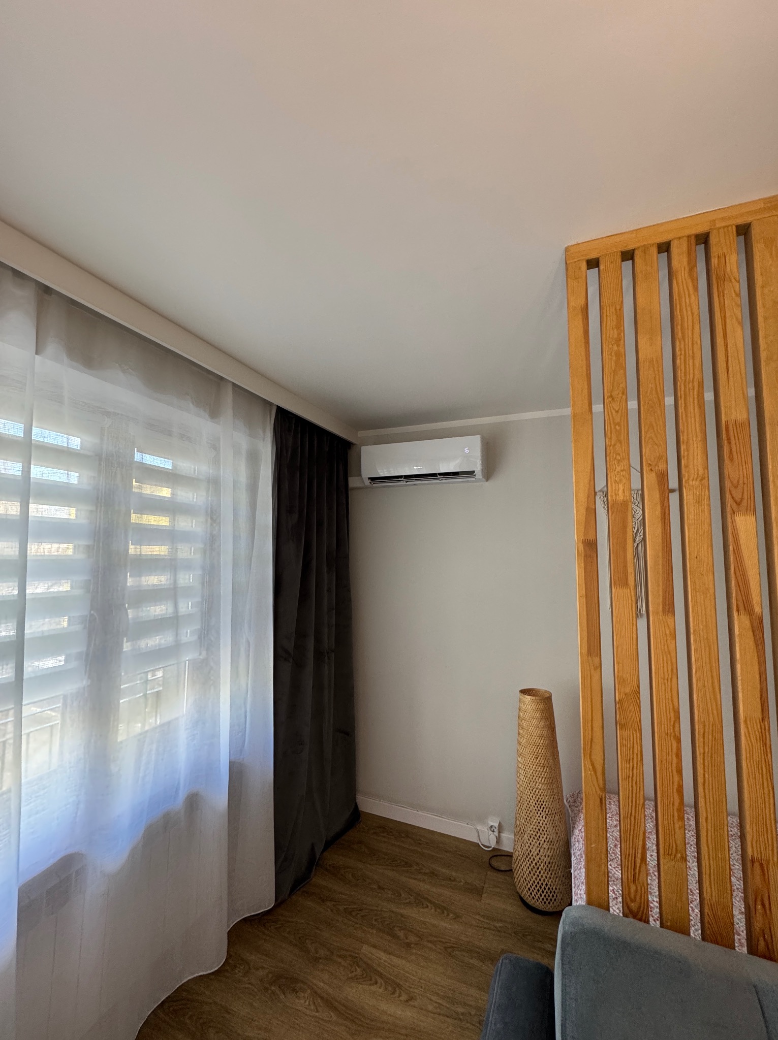 Biała klimatyzacja ścienna z wyświetlaczem temperatury, zamontowana w nowoczesnym salonie z drewnianą ścianką działową, zasłonami i lampą z wikliny.