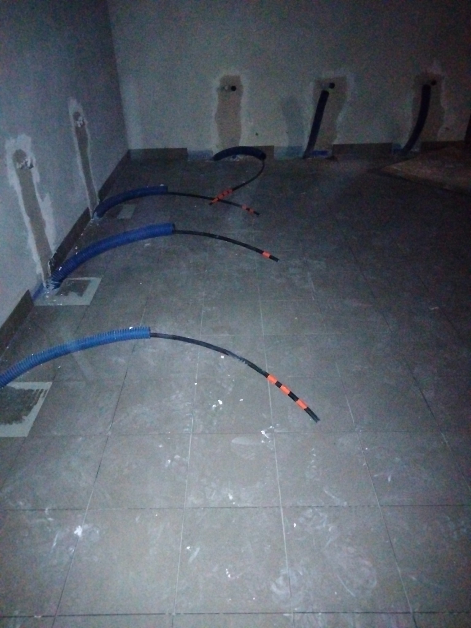 Wnętrze w trakcie instalacji elektrycznej: niebieskie rury karbowane prowadzą kable wzdłuż ściany, z otworami w ścianie, na szarych płytkach podłogowych widoczne ślady zaprawy.