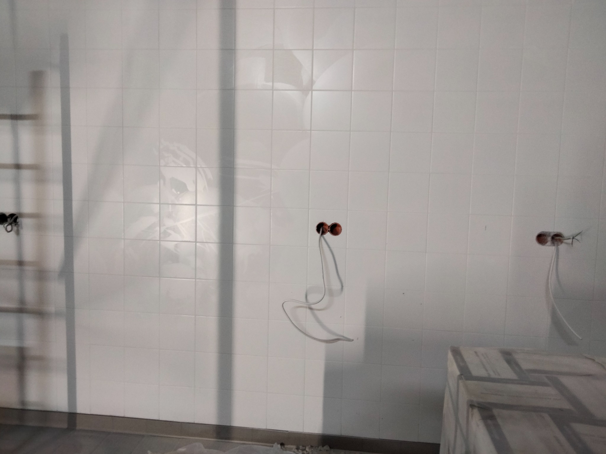 Ściana w łazience wyłożona białymi, błyszczącymi płytkami, widoczne przygotowane punkty elektryczne z wystającymi kablami oraz fragment drabiny i zabezpieczonego folią blatu.