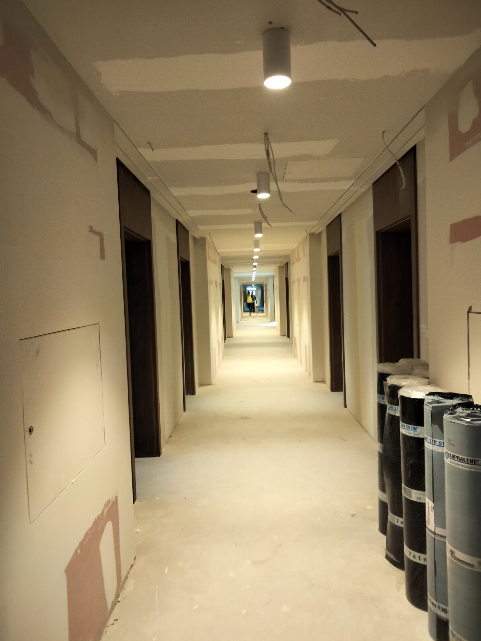 Długi korytarz w trakcie prac wykończeniowych, widoczne rolki papy na podłodze, ślady szpachlowania na ścianach i sufitach, oraz osoba w oddali w żółtej kamizelce.