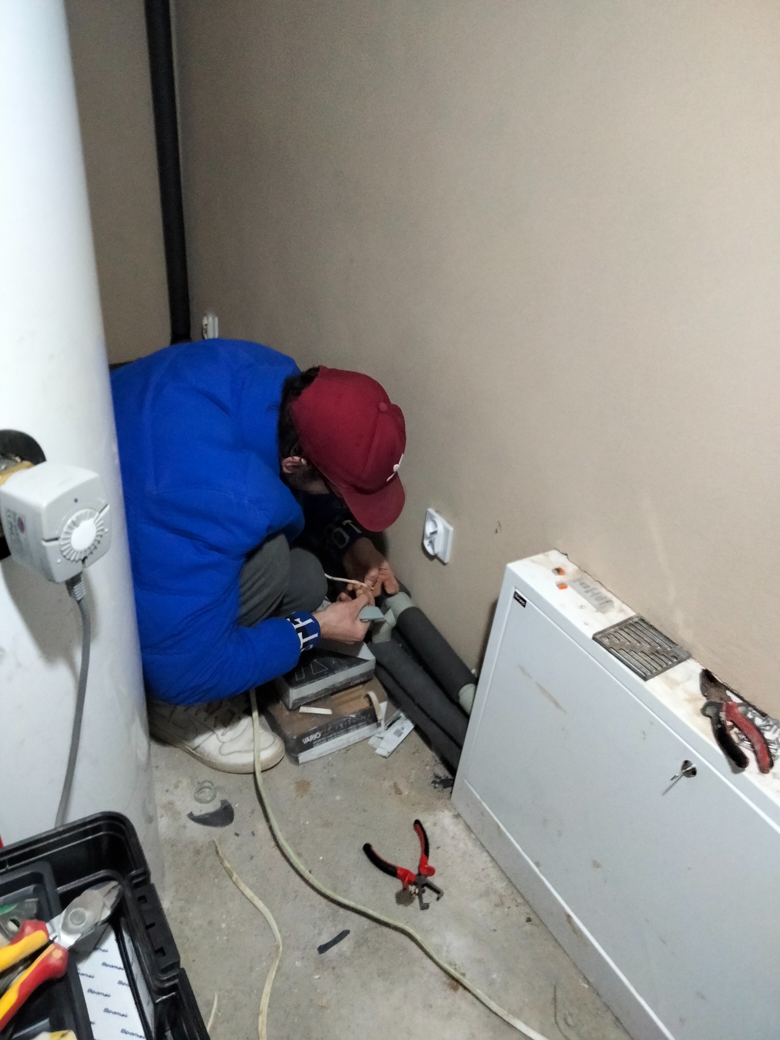 Elektryk w trakcie pracy, kuca przy białej skrzynce elektrycznej, w tle widoczny biały bojler, obok leżą narzędzia i kable