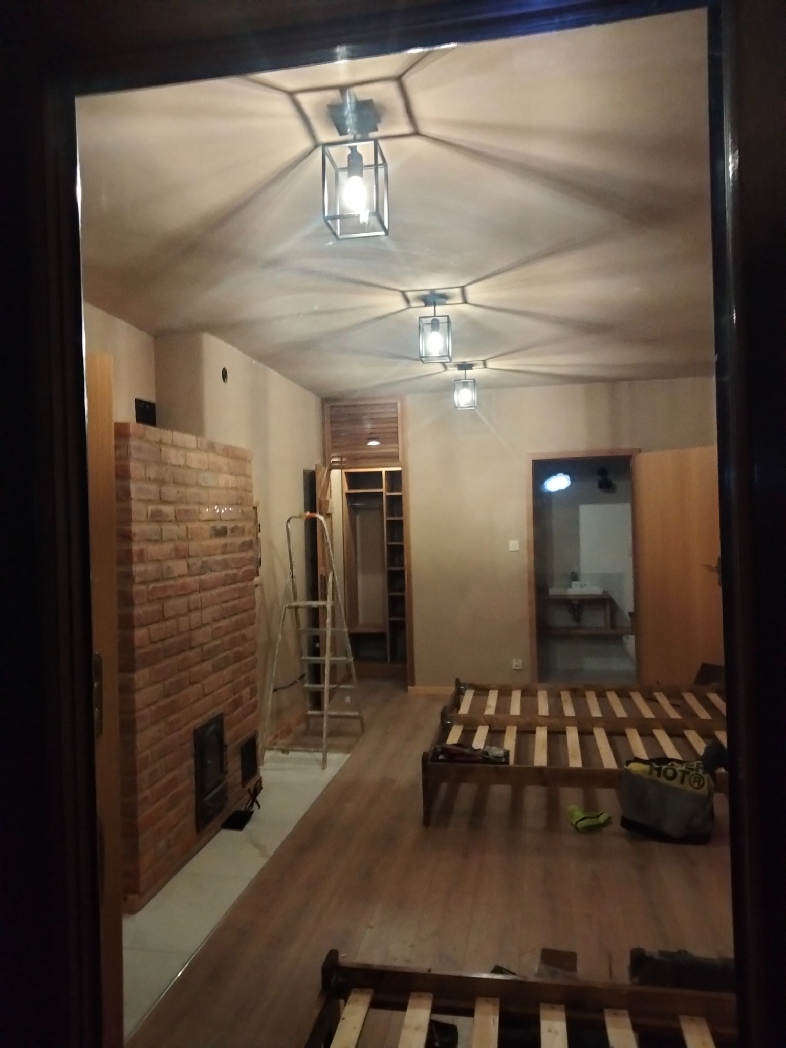 Wnętrze pokoju w trakcie remontu z widoczną ścianą z cegły, drabina oparta o ścianę, drewniane ramy łóżek na podłodze i nowoczesne lampy sufitowe z geometrycznymi kloszami.