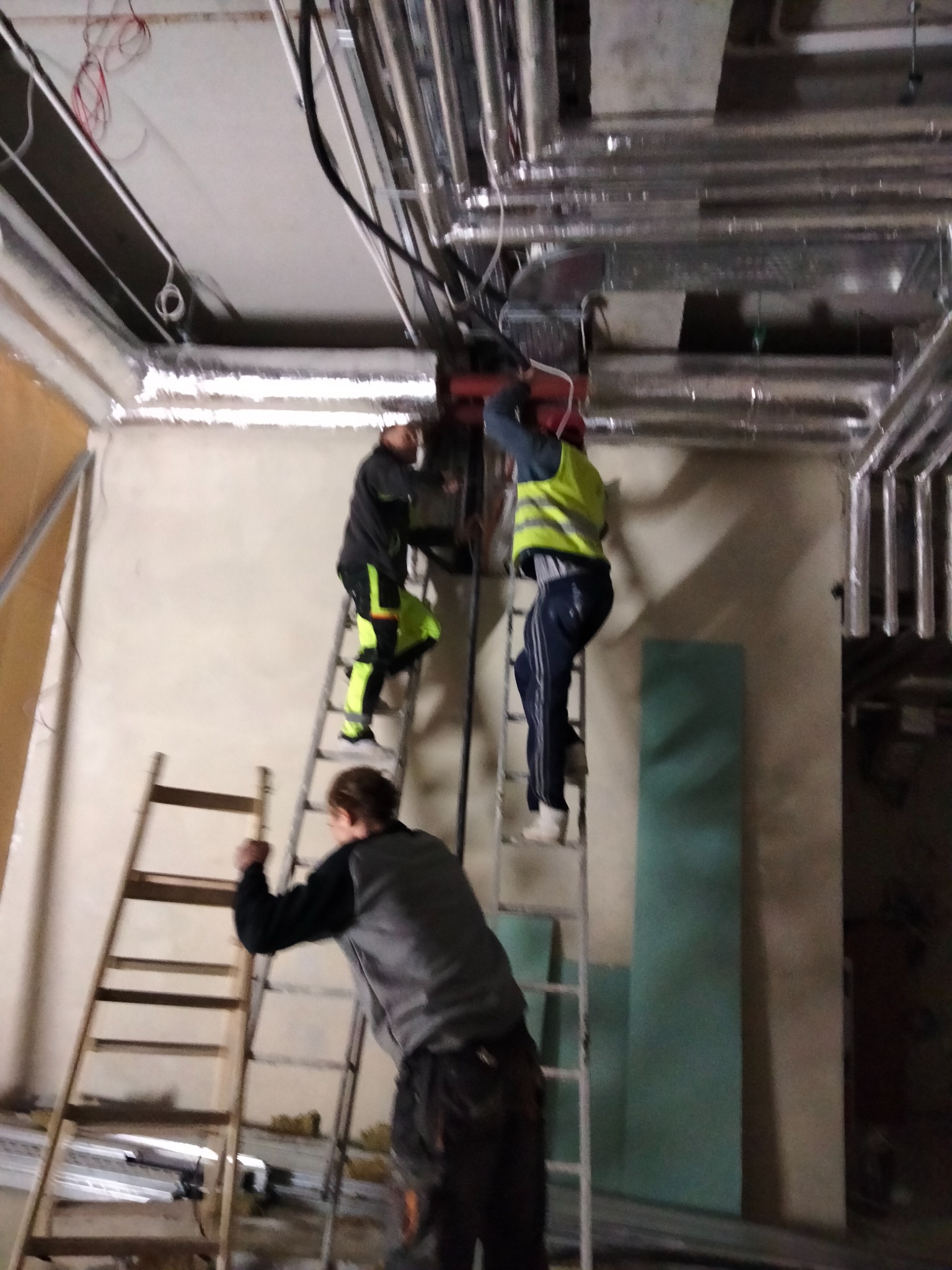 Trzech mężczyzn na drabinach instaluje rury i przewody pod sufitem w surowym wnętrzu. Widoczne rury wentylacyjne, kable elektryczne, ściany w stanie deweloperskim i płyty gipsowe.