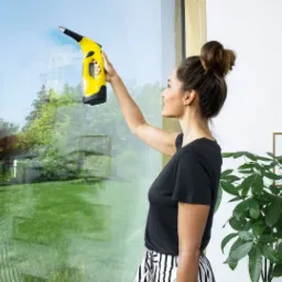 Kobieta z włosami upiętymi w kok używa elektrycznej myjki do okien marki Kärcher, widok na ogród przez umyte okno.
