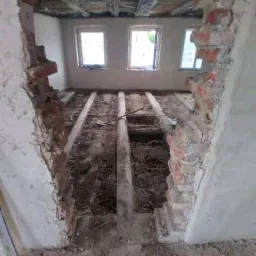 Wnętrze w trakcie rozbiórki: widoczne fragmenty muru po wyburzeniu ściany, odsłonięte belki stropowe i podłogowe, trzy okna w tle.