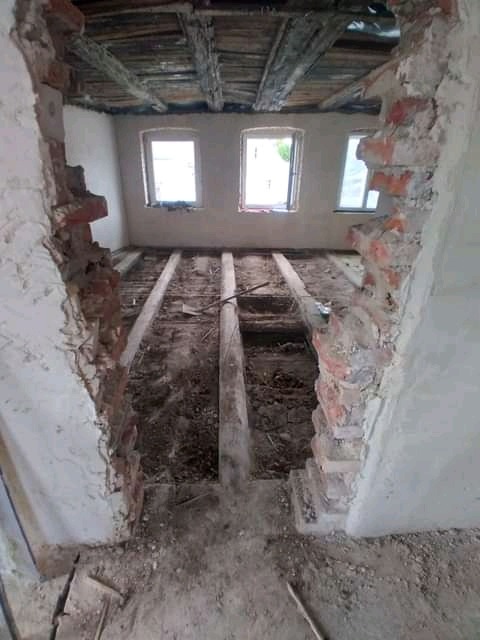 Wnętrze w trakcie rozbiórki: widoczne fragmenty muru po wyburzeniu ściany, odsłonięte belki stropowe i podłogowe, trzy okna w tle.