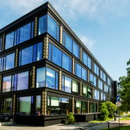 Nowoczesny, czterokondygnacyjny budynek biurowy z ciemną elewacją i dużymi, błękitnymi oknami w Warszawie, widziany z perspektywy chodnika. Na oknach widoczne odbicia otoczenia. Obok budynku drzewa...