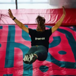 Kobieta w czarnej koszulce z napisem 'trener' skacze na trampolinie z wzorem w kolorze turkusowo-różowym.