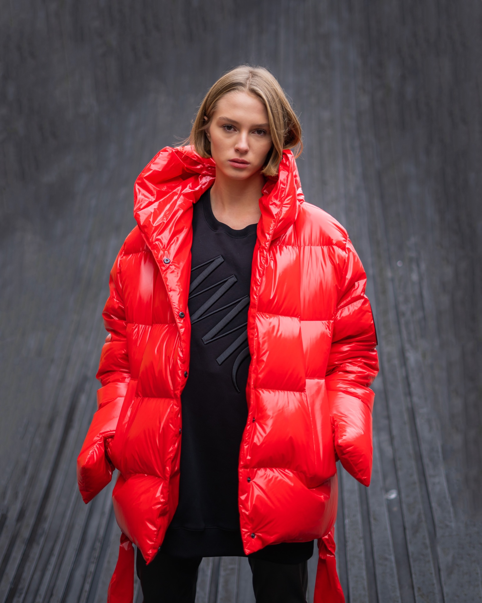 Kobieta w jaskrawoczerwonej, lakierowanej kurtce puchowej z kapturem, na tle szarej, teksturowanej ściany.