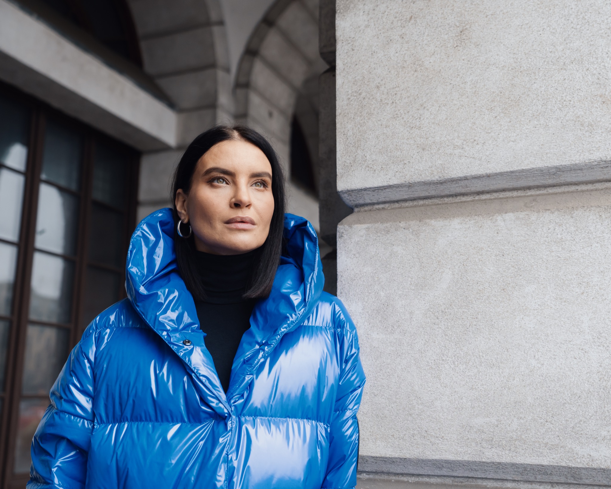 Portret kobiety w błyszczącej, niebieskiej kurtce puchowej na tle kamiennej fasady budynku z łukowymi przejściami.