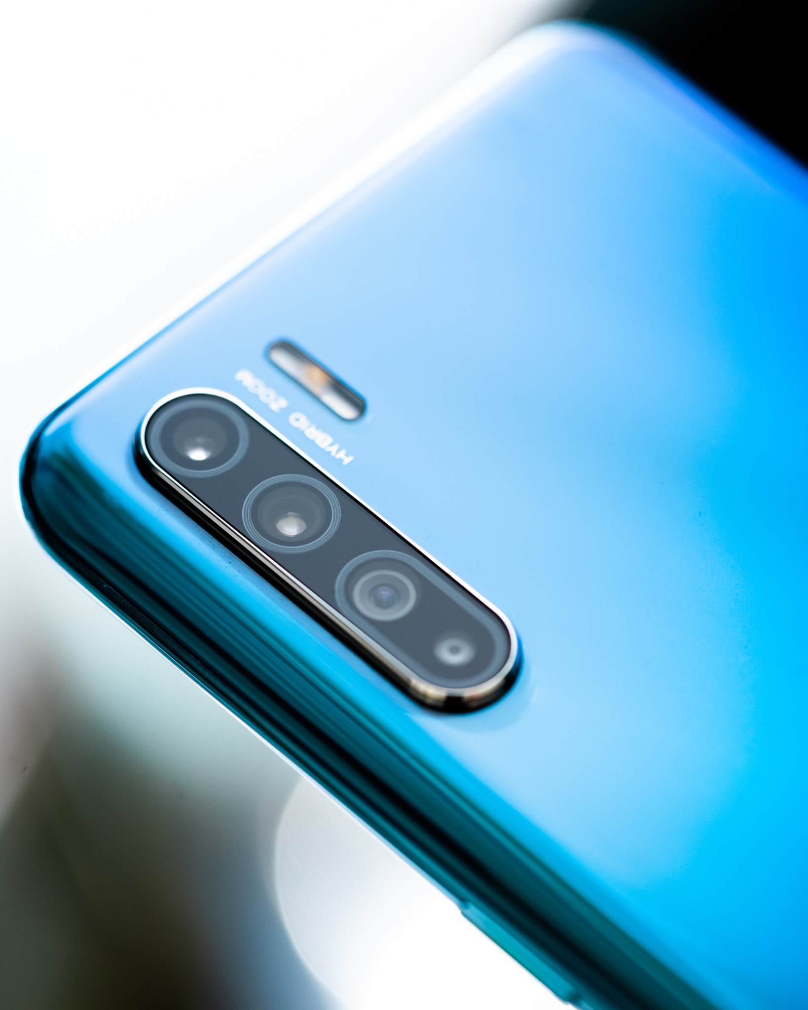 Błękitny smartfon z potrójnym aparatem i napisem 'Hybrid Zoom' na obudowie, leżący na jasnej, refleksyjnej powierzchni.