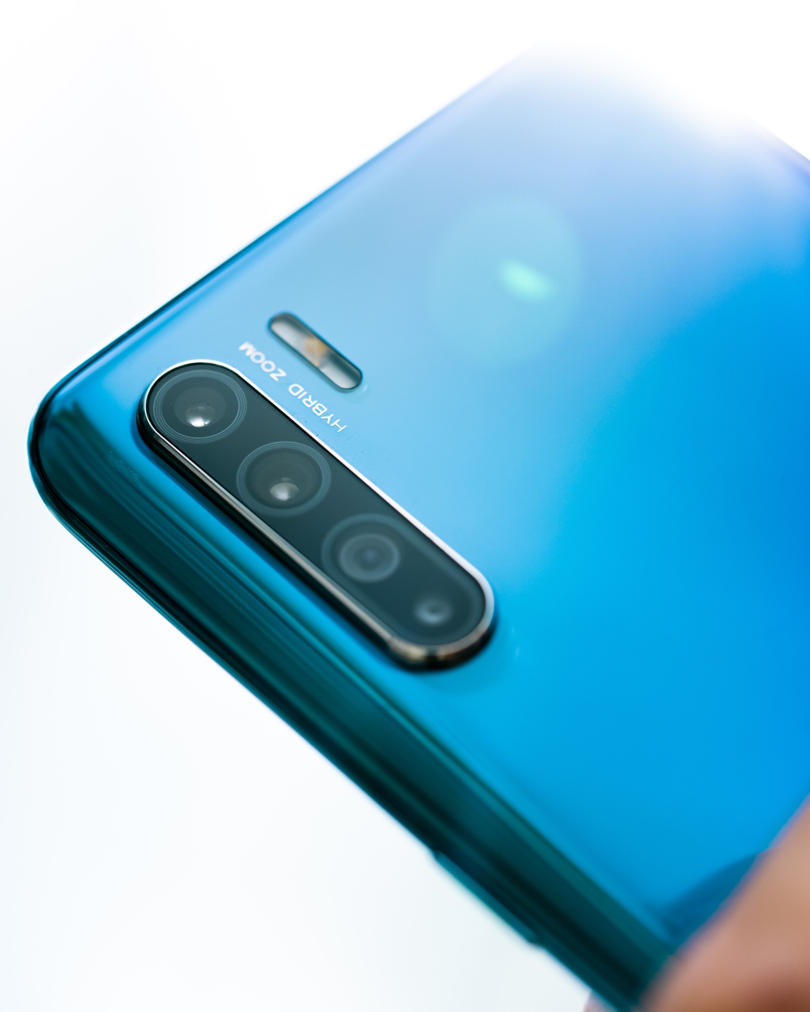 Zbliżenie na potrójny obiektyw aparatu w niebieskim smartfonie z napisem HYBRID ZOOM, skierowany pod kątem.